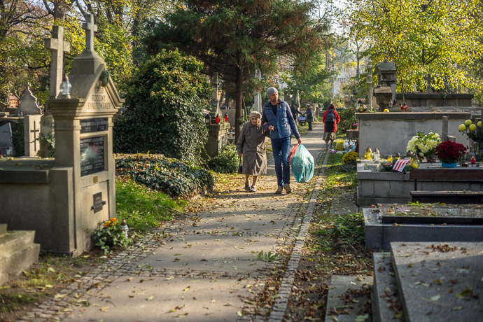A man supports an old woman and carries plastic bags full of flowers as they walk through the cemetery, All Saints' Day in  Image: Michel Meijer