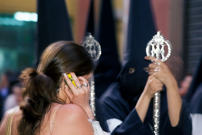 A hooded penitent stands in the sunshine waiting for the procession to progress, watching his girlfriend on the phone. Image: Michel Meijer