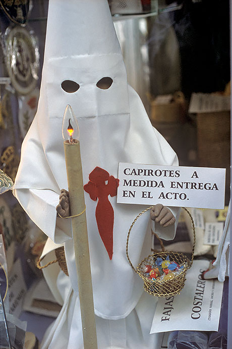 A doll, dressed as a hooded penitent, stands in a shop window and functions as an advertising figure. Image: Michel Meijer